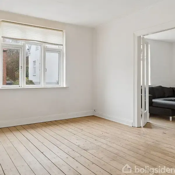 Et lyst værelse med trægulv og et stort vindue. Der er en åben dør til et andet rum, hvor en sort sofa kan ses.