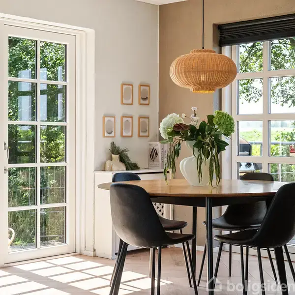Rundt spisebord med fire sorte stole står i lysfyldt rum. Hængende lampe over bordet. Store vinduer og dør mod have. Blomsterdekorationer på bordet og væggen. Ingen tekst.