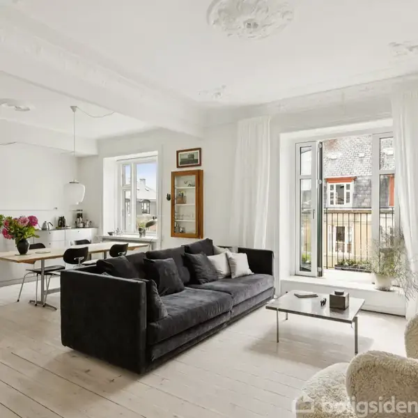 Stue med sort sofa vender mod et sofabord. I baggrunden ses et moderne køkken med spisebord og stole, vinduer giver udsigt til altan og lysindfald.