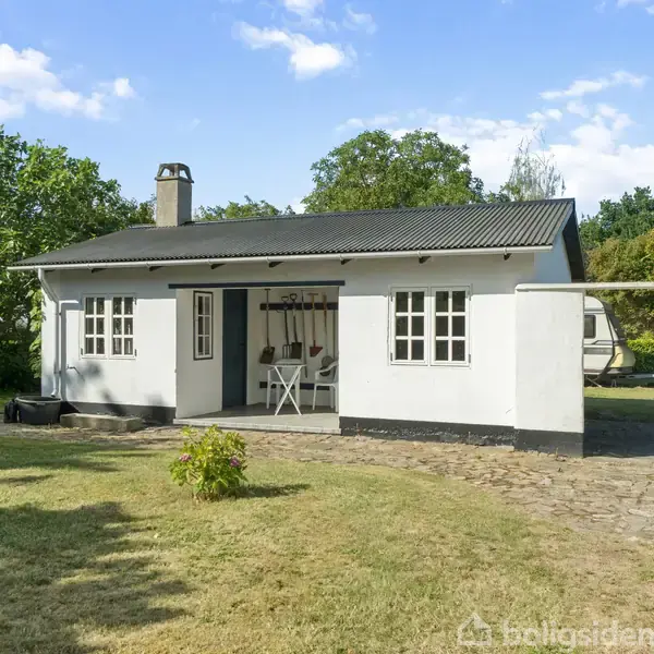 Et hvidt hus med sort tag står stille i en have med grønne træer og en græsplæne. Stenbelagte stier omgiver huset.