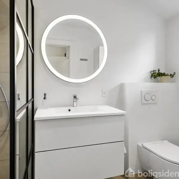 Et moderne badeværelse med en rund, oplyst spejl over en hvid vask. En bruser med glasdøre til venstre og et toilet til højre i et lyst rum.