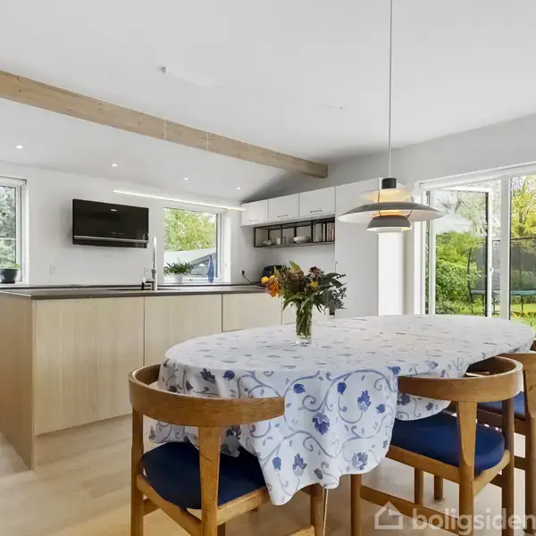 Et spisebord med blomster i vase står i et moderne køkken med lyst træ, store vinduer og udsigt til en grøn have. En lampe hænger over bordet.