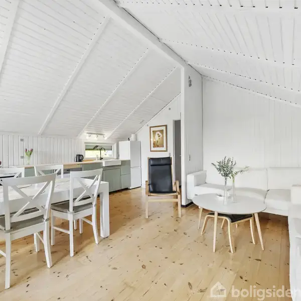 Et lyst, åbent rum med skrå lofter. En spiseplads med seks stole står ved siden af et sofahjørne med et hvidt bord. Køkkenet ses bagved, og der er dekorationer.