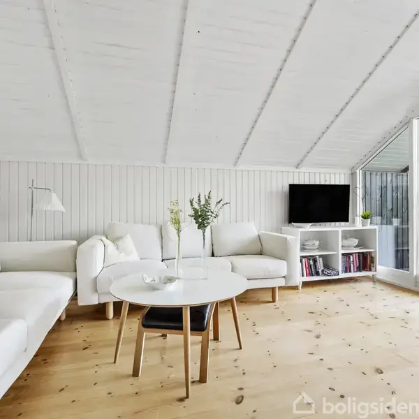 Stue med hvide sofaer arrangeret omkring et rundt sofabord. En fernisbelagt trægulv forbinder til en udendørs terrasse gennem store glasdøre. Et TV står på en hvid reol med bøger.