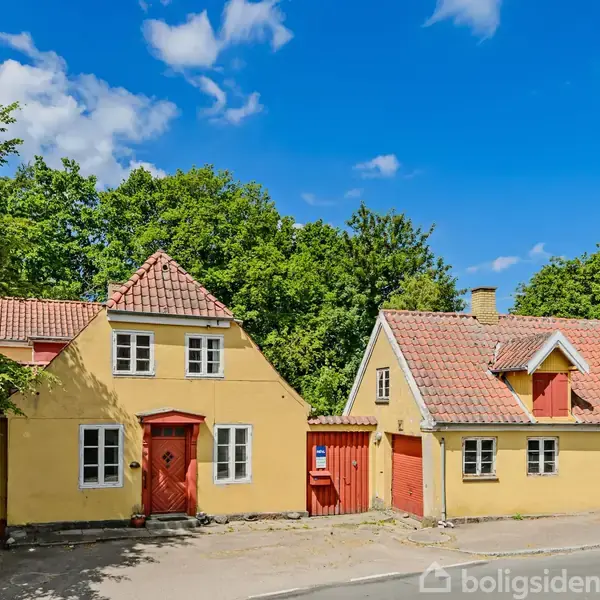 To gule, charmerende huse med røde teglstenstag står side om side ved en rolig vej. Grønne træer omkranser husene, og himlen er blå med få skyer.