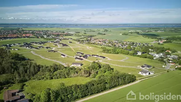Luftfoto over boligområde; moderne huse adskilt af veje omgivet af grønne marker og tæt på en landsby, udsigt over skov og vand i horisonten.