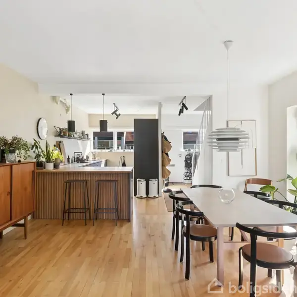 Spisebord med stole står i en moderne spisestue og køkken med trægulv og indbygget lys. Rummet er lyst med store vinduer, planter og skandinavisk design. Hylder og skuffer langs væggen.