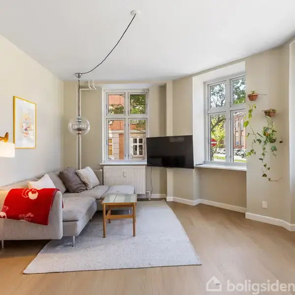 Sofa står i hjørne af stue dækket med tæppe og pude, under en væglampe. Stuen har trægulv, stort vindue, plante og vægmonteret TV, skaber lys og rummelig atmosfære.