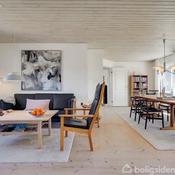Stue med sofa og lænestol omkring et sofabord; spisebord med stole i baggrunden. Væggene har kunstværker, og rummet får dagslys gennem store vinduer.