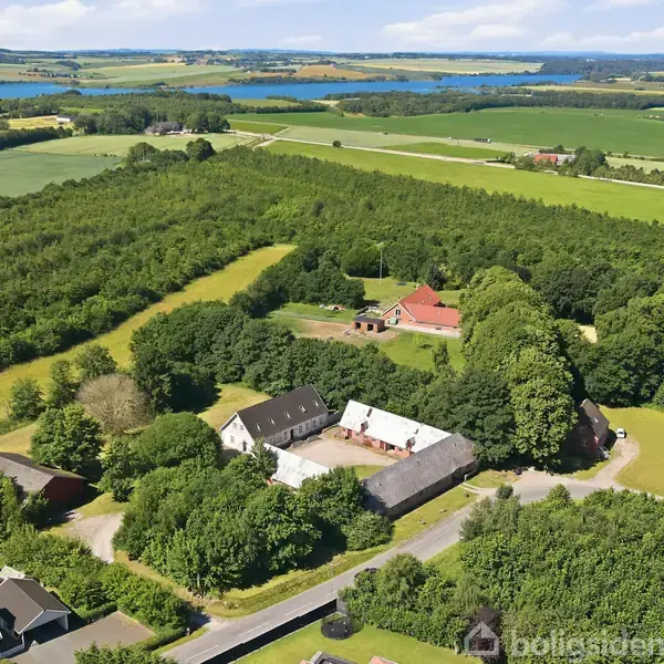 Gård omgivet af grønne marker og skov; bygningerne inkluderer en hovedbygning og lader. I baggrunden ses åbent landskab med spredte huse og en fjern sø.