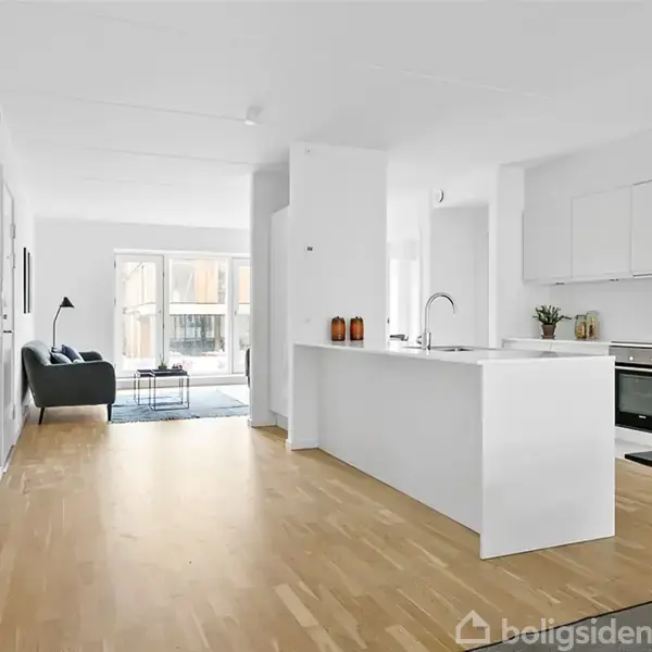 Et moderne køkken med hvidt interiør og ø, placeret i en lys stue med trægulv. En grå sofa og gulvlampe ses ved store vinduer, der bringer dagslys ind.