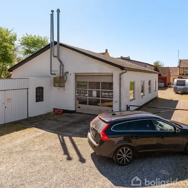 En sort bil holder parkeret på en grusplads foran en hvid bygning. Bygningen har et skilt med teksten "CYKLER" og "Årets bil" på ruden. Baggrunden viser en vej og flere bygninger.