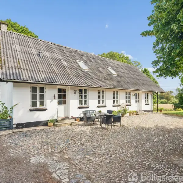 Hvidt bondehus med skråt tag, omgivet af grusbelagt gårdsplads og frodige træer. Der står havemøbler foran huset, som skaber en hyggelig udendørs atmosfære i landlige omgivelser.
