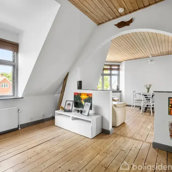 Et lyst stueområde med trægulv og hvide vægge viser en fladskærm på en tv-bænk. Spisebord og stole er synlige i baggrunden under et loft med træpaneler.