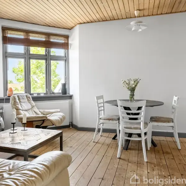 Et lyst stuehjørne med en rund spisebord med fire stole, en lænestol ved vinduet, trægulv og træloft. En vinduesudsigt viser grønne træer udenfor.
