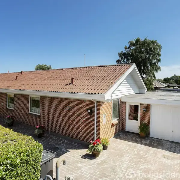 Murstenshus med rød tegltag står stille i sollys, omkranset af smuk, grøn hæk og flisebelagt indkørsel. Til højre ses en hvid garage.