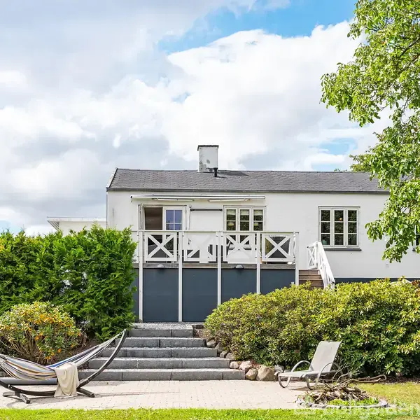 Et hvidt hus med en balkon, placeret i en have med grønne buske og store træer. Foran huset er der en hængekøje og en solstol på en flisebelagt terrasse.