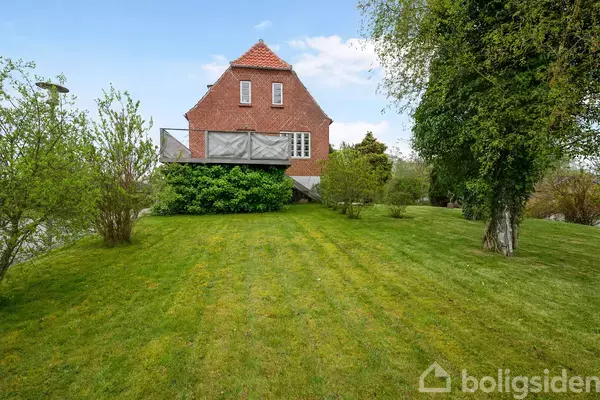 En rød murstensbygning med en altan er omgivet af en frodig have med græsplæne og træer. Solrig himmel udenfor ses i baggrunden.