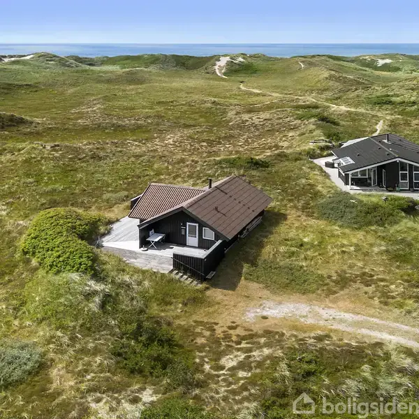 En sortmalet træhytte med skråt tag står stationær på en grøn klitlandskab med nogle få andre huse i nærheden og havet i horisonten.