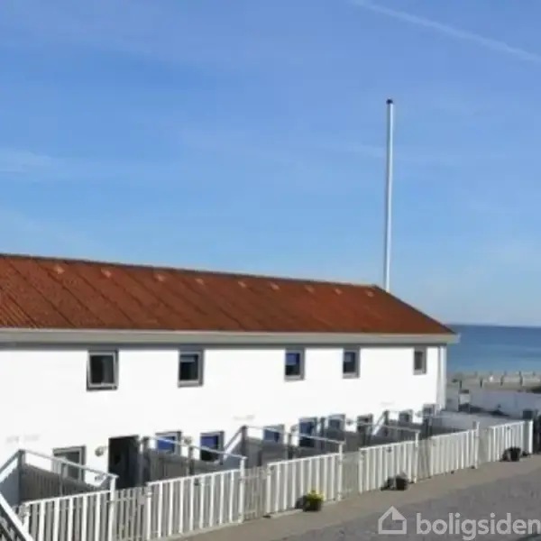 En hvid bygning med rødt tegltag står stille ved kysten, omgivet af hegn og med havudsigt i baggrunden under en blå himmel.