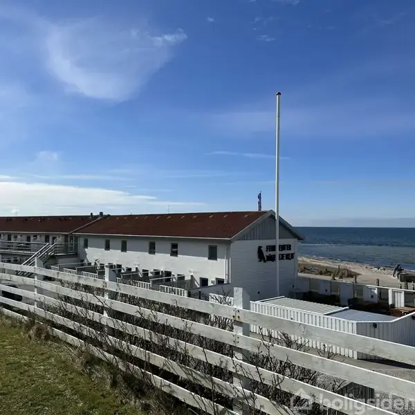 En hvid bygning med rødt tag står langs en strandpromenade på en lysdag med havet i baggrunden. En hvid træhegn omkranser bygningen.