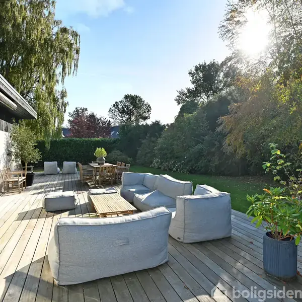 Terrasse med polstrede møbler og træbord, der står på trægulv. Sollys skinner over en have fyldt med træer og buske.