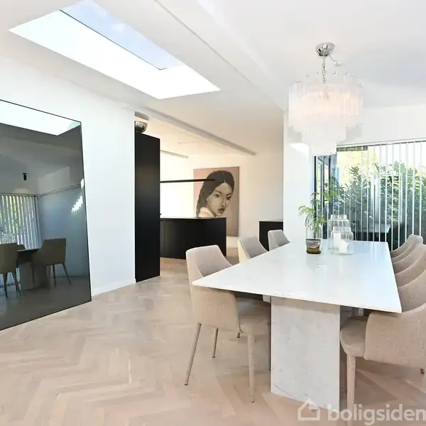 Et moderne spisebord med syv beige stole står i et lyst rum med parketgulv. Store spejle og planteliv omkranser, mens et stort portræt hænger ved siden af et ovenlysvindue.