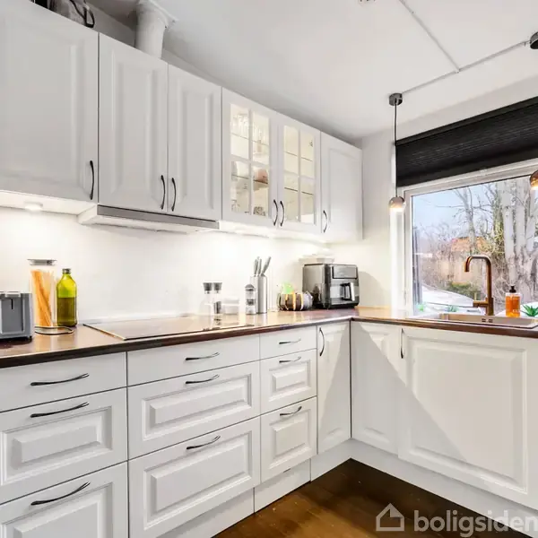 Et moderne, lyst køkken med hvide skabe og sort bordplade. Bordpladen er udstyret med en kaffemaskine, knivholder og flasker. Et stort vindue giver udsigt til en have.