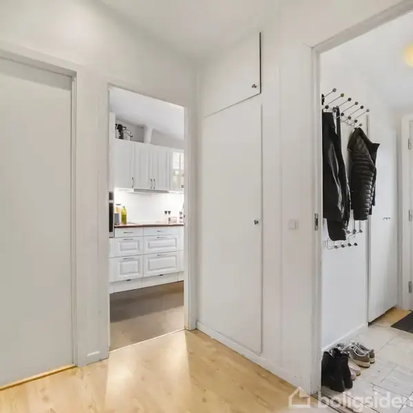 En entré med lyst trægulv har jakker hængende på knager. Til venstre ses en lukket dør, og mod højre er en dør til køkkenet med hvide skabe.