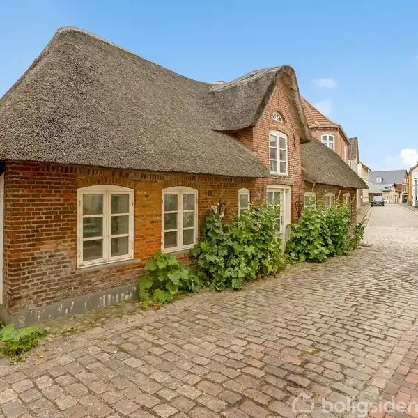 Stråtækt bindingsværkshus med små vinduer, omgivet af brostensbelagt gade og andre traditionelle bygninger. Grønne planter vokser op ad murene, og himlen er klar og blå.