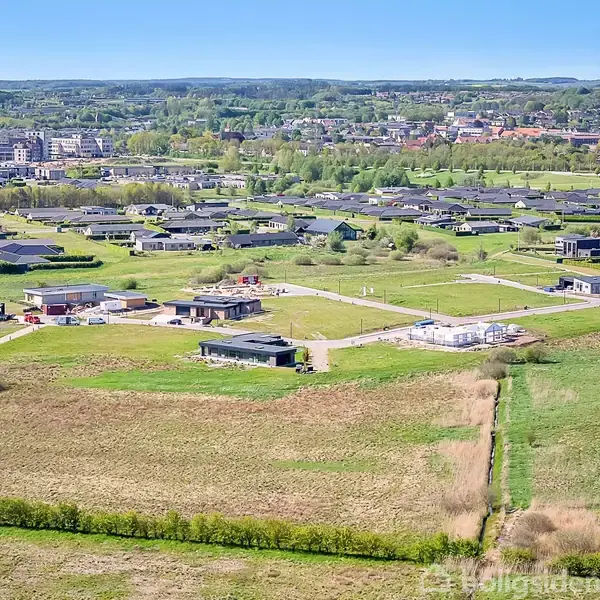 Boliger er placeret i et vidtstrakt landskabsområde. Huse med flade tage står spredt langs snoede veje. Omkringliggende marker og skove omkranser bebyggelsen samt en fjern by i baggrunden.