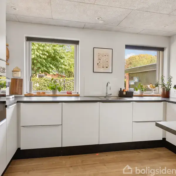 Hvidt køkken med vask integreret i sort bordplade. Vinduer med udsigt til grønne træer og planter på vindueskarmen. Moderne indretning i lys, minimalistisk stil.