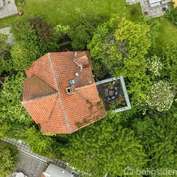 Et hus med rødt tag ses fra oven. Huset er omgivet af frodige grønne træer og en have. Der er en terrasse med havemøbler ved siden af huset.