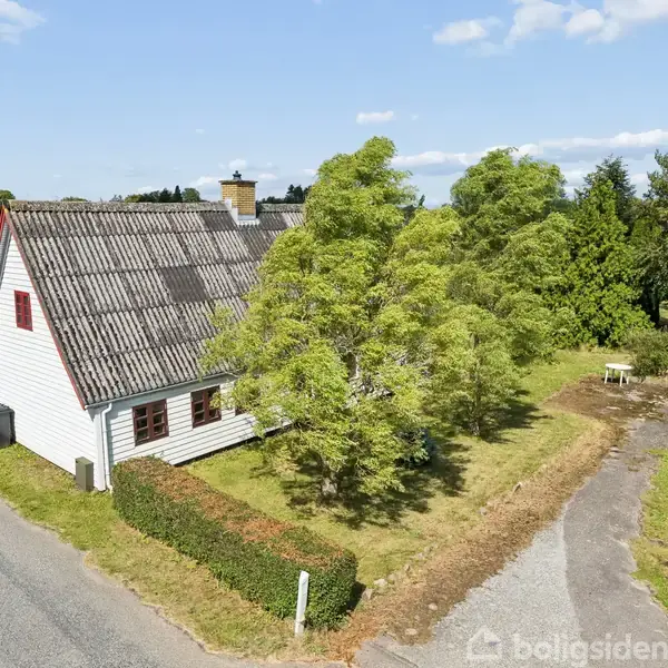 Hus med rødt tag og hvide vægge står ved vej; omgivet af træer og grønt område. Skyfri himmel og solrig dag.