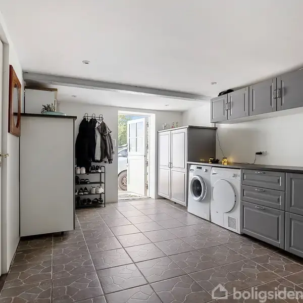 Vaskemaskine og skab står langs en væg med grå skabe. På gulvet er der fliser, mens døren leder ud til en have. Jakker hænger til venstre på en knagerække.