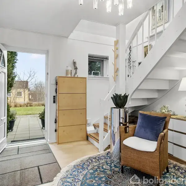 En åben hoveddør fører til en lys entre med trappe til højre, en kurvestol placeret på et blåt orientalsk tæppe, og en kommode med planter og dekorationer.