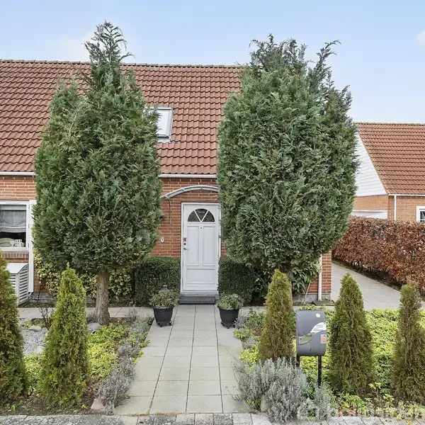 Hus med rødder tag ses forfra. En hvid dør omkranset af to høje, grønne træer. Foran er der en række mindre buske langs en flisebelagt sti.