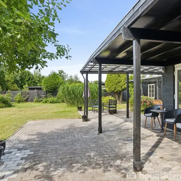 En overdækket terrasse med sorte søjler står foran et hus i en have. Der er havemøbler under taget, og i baggrunden ses en grøn græsplæne med træer og buske.