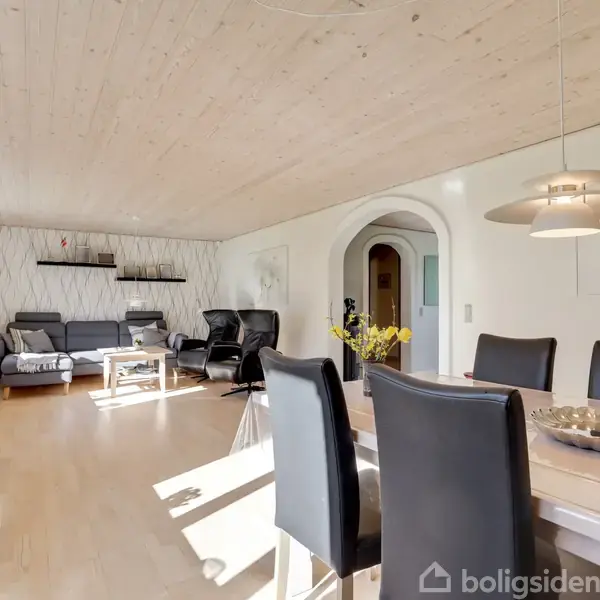 Langbord dækket med skål står i fornemt spisestue med sorte stole. I baggrunden afslappede sofaer, TV på væggen, åbning til gang og dekoreret vægmaleri.
