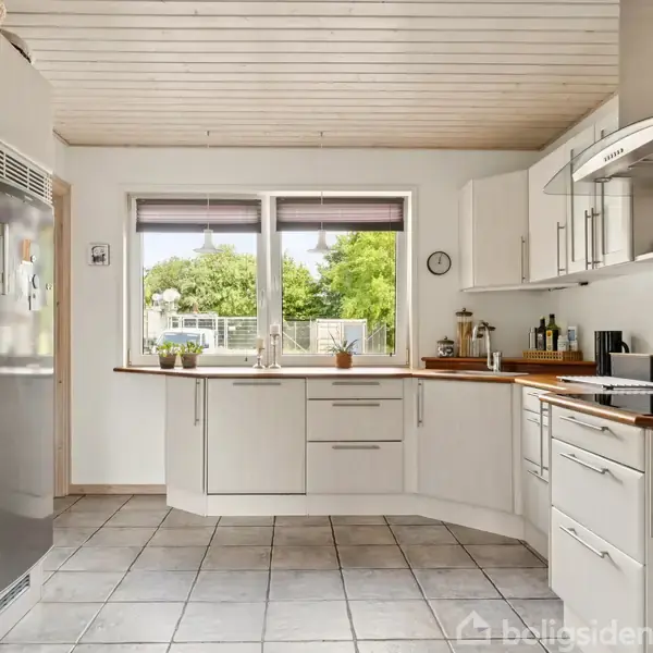 Et moderne køkken med hvidt interiør står i ro. Rustfrit stål og træoverflader dominerer. Store vinduer giver udsigt til en grøn have. Flisegulv og loft med træplanker.