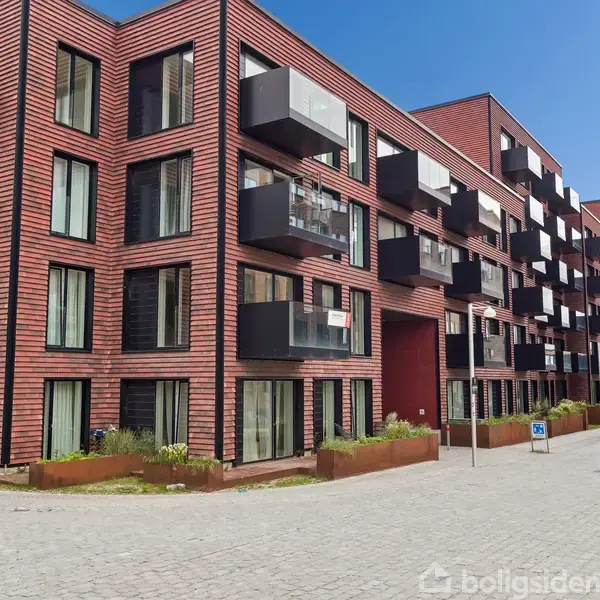 Lejlighedskompleks med røde mursten og sorte altaner placeret langs en brolagt gade. Bygningen har flere etager med store vinduer. Blå himmel i baggrunden uden synlig tekst.