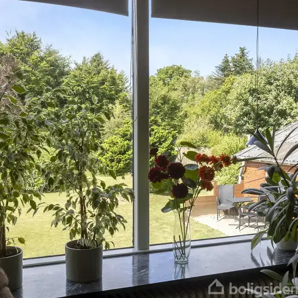 Planter i potter og en blomstervase står på vindueskarmen; udenfor ses en frodig have med buske, træer og en terrasse med møbler.