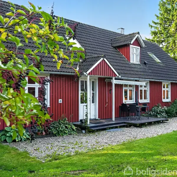 Et rødt træhus med hvide vinduesrammer står i en frodig have med grusvej. Træer og buske omgiver huset, som har en lille terrasse ved indgangen.