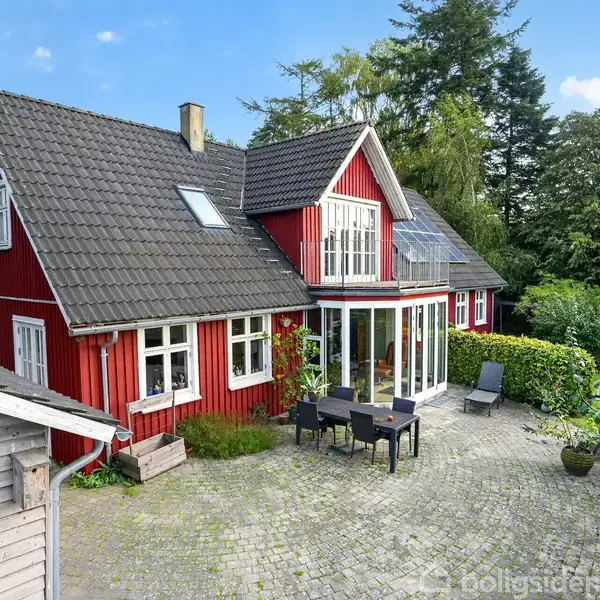 Rødt hus med skråtag står stille omgivet af grønne træer. Husets gårdhave har havemøbler, og fliser dækker jorden. En solrig dag skaber en lys og hyggelig atmosfære.