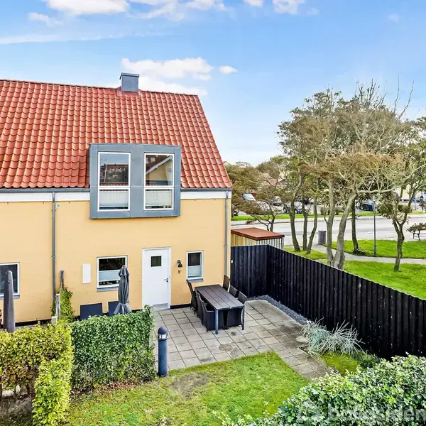 Et to-etagers rækkehus med rød tagsten og lyse mure, der står i en grøn have med patio og sorte stakit mod en vej med træer og græsareal.