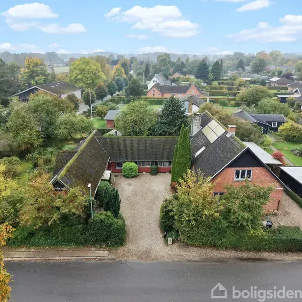 Et murstenshus med skråt tag ligger i en frodig have omkranset af træer og buske. En grusvej fører hen til hovedindgangen fra en stille villavej.