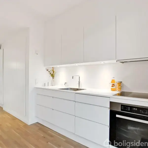 Hvide køkkenskabe står langs en væg; en vask og emhætte er indbygget, mens appelsinjuice kartoner er på bordpladen. Trægulve strækker sig mod en lys stue med store vinduer.