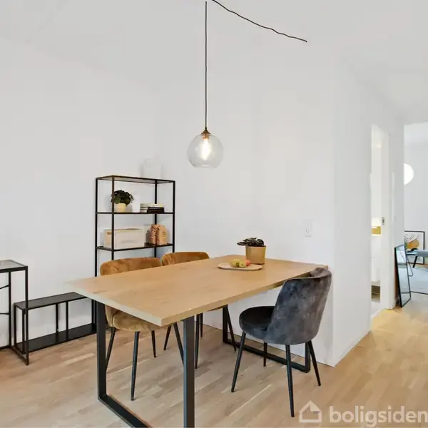Et træbord med tre stole omkring står i en lys, minimalistisk spisestue. En hylde med bøger og planter er i baggrunden, og et opholdsrum ses via en døråbning.