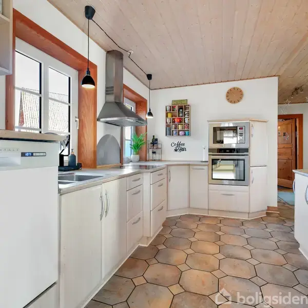 Et hvidt køkken med moderne apparater, som en ovn og mikrobølgeovn, placeret i et veloplyst rum med flisegulv. På væggen hænger dekorationer og et ur over en hvid bordplade.