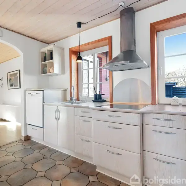 Et lyst køkken med hvide skabe, en grå emhætte og et spisebord i baggrunden. Vinduerne giver udsigt til en have. Text: "Carpe diem".